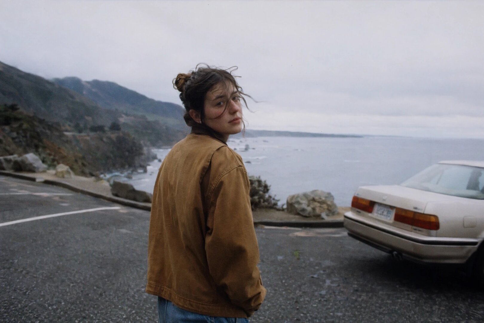 „A photorealistic candid travel scene of a person standing at a coastal roadside turnout on an overcast morning, shot on 35mm film. Natural imperfect framing, visible grain, ambient light, muted colors, wind in clothing and hair, cinematic realism, and the feeling of a lived-in documentary photograph.“